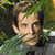 Pixelated image of a person with light brown hair and green foliage on the head, wearing a green and gray garment against a blurred green background.