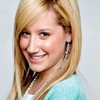 A person with long blonde hair wearing a light blue cardigan over a white top, smiling with visible teeth against a light gray background.