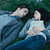 Two figures lie side-by-side in water, one with dark hair and blue clothing, the other with light-colored hair and a white top against a greenish background.