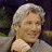 A pixelated image of a person with light-colored hair in a side ponytail, wearing a light shirt against a dark background.