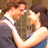 A couple in formal attire embraces and kisses, with the woman wearing a blue dress and the man in a dark suit.