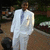 A person in a white suit stands on a paved path with greenery and blurred figures in the background, captured in a static image with no visible motion.