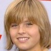 A static close-up of a person with light brown hair and bangs, smiling with visible teeth against a light blue background.