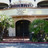 Static pixelated image of a building facade with dark doorway, greenery, and light-colored walls under a bright sky.