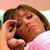 A person with brown hair and pink clothing holds a phone to their ear, making a heart shape with one hand while resting on a light green surface.