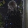 Animated sequence of a person with blonde hair in a dark coat standing near a with greenery, showing subtle head and body movements