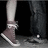 Pixelated close-up of a person's lower legs and feet wearing light gray pants and brown shoes with white soles against a dark background.