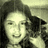 A pixelated grayscale image of a person with long dark hair, wearing a light-colored top and holding an object near their face.
