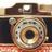 A pixelated close-up of a vintage-style camera with a prominent lens and flash, rendered in muted brown, beige, and gray tones.