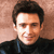Pixelated close-up of a person with dark hair wearing a dark high-collared shirt against a light brown background.