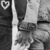 Pixelated grayscale image of a figure in dark pants and light shirt standing near a chair with visible legs, left side shows partial heart symbol.