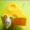 A white mouse with black eyes and a pink nose stands in front of a large yellow cheese wedge with orange rind on a green background.