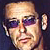 Pixelated static icon of a person with short light hair wearing dark sunglasses and an earpiece, set against a dark background.