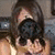 A person with brown hair and a light-colored sleeveless top holds a dark object near their face, positioned against a blurred background.