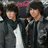 Two individuals in dark jackets stand side-by-side against a blurred background with blue text reading "I Love Nick & Joe" on the right.