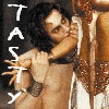 A person with long dark hair and a white forehead marking embraces another person in a sparkly outfit, with vertical text reading "TASTY" on the left side.