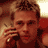 Pixelated static image of a man with short brown hair holding a microphone near his mouth, wearing a dark red sleeveless shirt against a warm-toned background.
