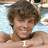 A person with brown curly hair lies on a light blue patterned surface, smiling at the camera with text overlays changing from 'I' to '<3' to 'Ryan'.