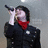 A person in a black jacket and red scarf holds a microphone near their mouth against a light gray background.