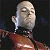 Pixelated static image of a bald cartoon character with closed eyes and a neutral expression, wearing a dark shirt over brown pants against a black background.