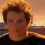 Pixelated image of a person with light brown curly hair wearing dark clothing against an outdoor background with buildings and sky.
