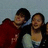 Two people pose together in a pixelated image with red text overlays on a black background, one in a red top and the other in a white top with dark hair.