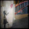 A grayscale silhouette of a girl holding a red heart-shaped balloon against a textured wall with a blurred background of a building entrance.