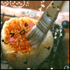 A round orange-brown food item with a smiling face is being brushed with a paintbrush, surrounded by small blue and pink decorative elements.
