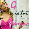 A woman in a pink lace top and white cardigan stands against a tiled wall with text overlay reading 'G is for glamour' in pink and black lettering.