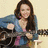 A woman with long brown hair in a blue denim jacket over pink clothing holds an electric guitar against a light background.