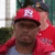 Pixelated image of a person wearing a red cap with a white 'R' symbol and red clothing, set against a blurred background with green and white elements.