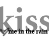Static grayscale icon featuring large stylized text 'kiss' with smaller black text 'me in the rain' below, including a small checkmark symbol on the left side of the lower text.