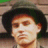 Pixelated close-up of a person with dark hair and light skin, wearing a dark hat, against a blurred background with green and brown tones.
