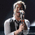 A person with light-colored hair holds a microphone, wearing a white shirt and dark vest against a dark background.