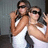 Two people in white tank tops pose closely together, one holding a camera to their face against a dark background.