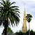 Pixelated static image shows palm trees and a tall tower against a light blue sky with green foliage at the base.