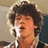 Pixelated static image of a person with dark curly hair wearing a white shirt and dark jacket, mouth open in mid-expression against a muted background.
