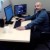Pixelated static image shows a person in dark clothing seated at a desk with two computer monitors, one blue and one dark blue, on a light-colored surface.