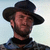 Static pixelated image of a man wearing a dark wide-brimmed hat and dark clothing against a light background, with visible facial features including beard stubble and earlobe.