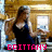 Pixelated image of a person with long brown hair wearing a black sleeveless top, standing in an indoor setting with wooden walls and a window.