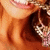 A close-up of a smiling person with light brown hair and a gold chain necklace, showing a partial view of the face and neck.