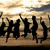 Silhouettes of multiple figures jumping against a sunset sky with clouds and sandy ground.