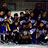 Group of people in blue and white uniforms with yellow sashes, seated together under a dark background.