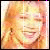 A static image of a person with light brown hair and bangs, wearing earrings, smiling broadly against a plain background.