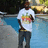 Pixelated static image of a person standing outdoors wearing a white t-shirt with colorful graphic, dark pants, and carrying a brown strap across the chest.