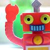 Red robot with yellow eyes and blue teeth waves right arm, green background, yellow circular chest emblem.