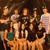 A pixelated group of figures in warm tones with a dark background, featuring multiple human-like forms in varied poses and clothing.