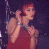 A person with short red hair performs on stage, wearing a sleeveless red top and dark vest, holding a microphone in various poses.