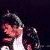 A person with dark hair and a white shirt appears in , wearing a black jacket with silver zippers against a dark background