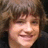 Pixelated close-up of a smiling person with brown hair and light skin, cropped to show face and upper shoulders against a dark background.